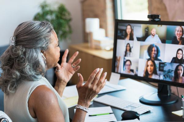 A woman with white hair and dark skin facing a computer screen that has faces of people in a remote meeting.